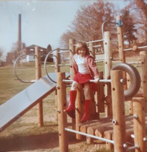 Sara Ryan as a small kid on a playground structure including a tire and a slide, wearing a red hoodie, white t-shirt, red skirt and bright red knee-high boots.