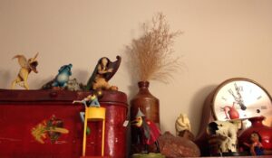 Several Hieronymus Bosch figurines on top of a bookcase, plus an old red metal box, a Steiff hedgehog, a Chris Ware "Super-man" figurine, an ink bottle with dried flowers, a seashell, a small red vase, and a stopped clock with Roman numerals. 